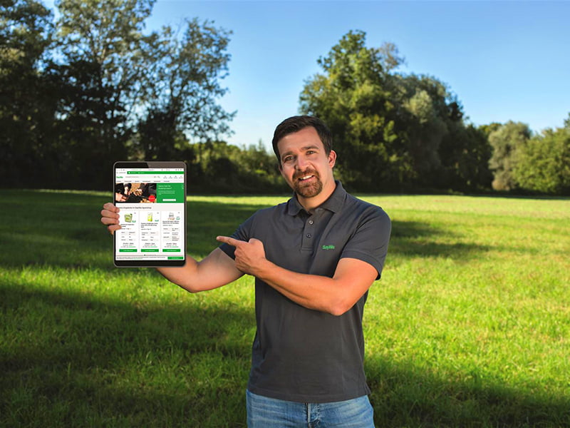 Mann steht in einer Wiese und hält in einer Hand das Tablet und mit der anderen Hand zeigt er auf das Tablet.