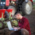 Fiatal gazdálkodó laptoppal térdel a vetőgép mögött