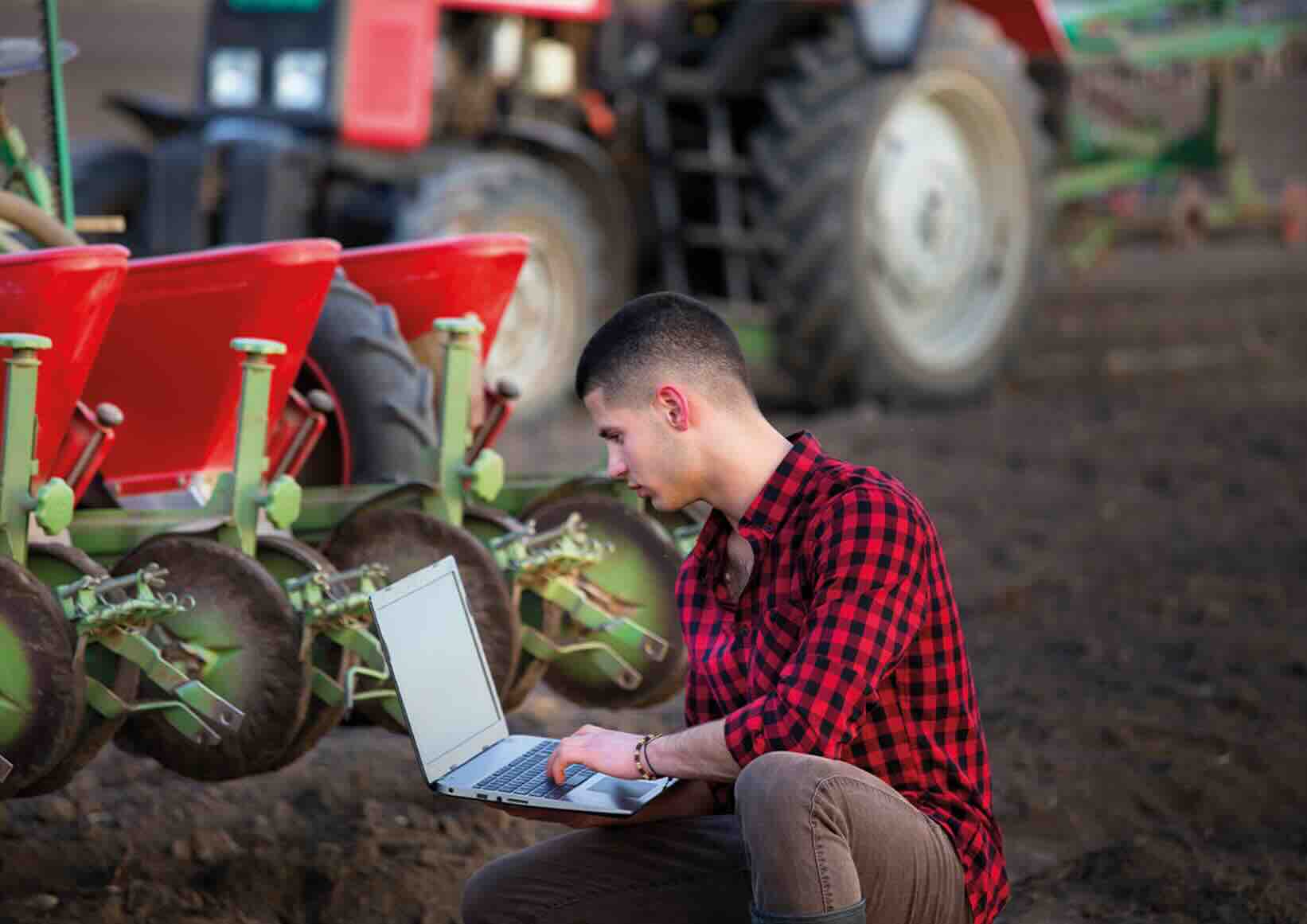 Fiatal gazdálkodó laptoppal térdel a vetőgép mögött